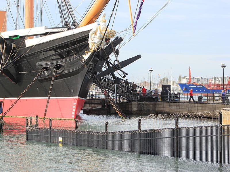 Black security fencing with barbed wire in sea at Portsmouth Historic Dockyard with historic ship behind